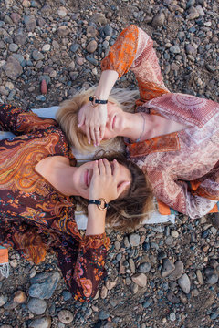 Overhead view of two women lying on pebbles, covering eyes
