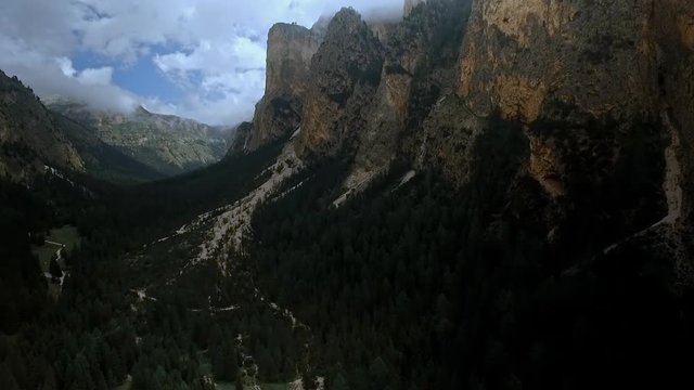 Steady aerial shot with swirling clouds above a dark, imposing and rugged valley with high cliffs and dark forests.