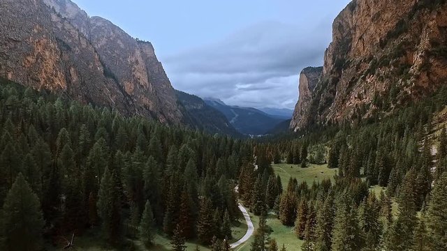 Aerial of a drone crossing a beautiful wooded valley in the Alps. Towering cliffs and dark green spruce trees. A hiking path snakes through the landscape.