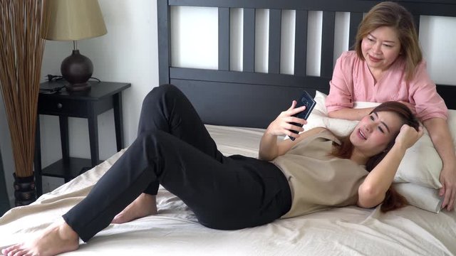 4K Wide shot of Happy relaxation beautiful young adult asian daughter lying down on the  bed with her senior retired mother in bed room and using smartphone with internet and social media together.
