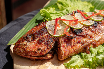 Fried fish with fresh green salad and lemon of Bali, Indonesia, closeup. Delicious roasted sea fish with lemon on wooden plate in restaurant