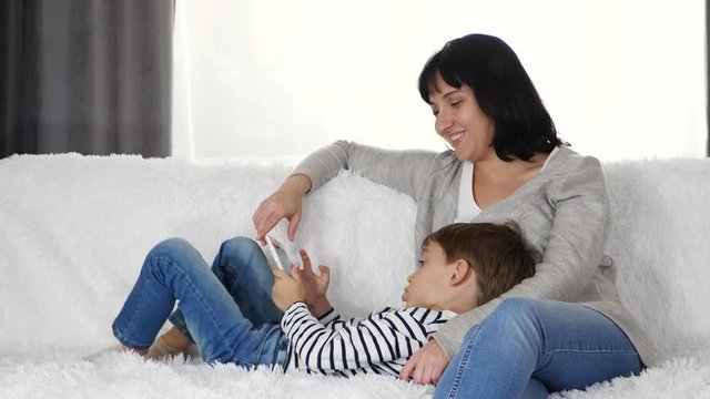 Happy family: mother and son spend time playing with a tablet. Woman and child sitting on the sofa. The concept of raising children using technology.