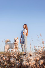 Obraz premium Girl on a walk with her dog. She holds a dog on a leash. The girl is dressed in a blue denim overalls. Dog breed Samoyed husky.