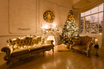 Christmas evening by candlelight. classic apartments with a white fireplace, decorated tree, sofa, large windows and chandelier.