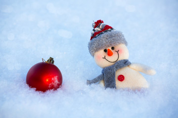 There is a happy funny toy snowman standing on the snow and a red glass ball. The snowman is wearing a fur hat and scarf. The concept of winter, Christmas, new year. Copy space. horizontal orientation