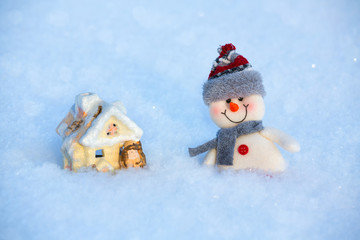 On the snow is a happy funny toy snowman and toy house. The snowman is wearing a fur hat and scarf. The concept of winter, Christmas, New year. Copy space. horizontal orientation.