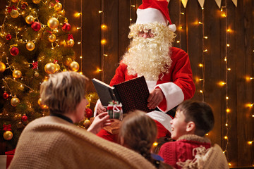 Fototapeta na wymiar Santa Claus reading book for family. Mother and children sitting indoor near decorated xmas tree with lights - Merry Christmas and Happy Holidays!