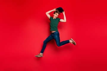 Full length profile side photo of cheerful crazy guy jump run have fun hold his blue headwear wear casual style green t-shirt sneakers isolated over red color background