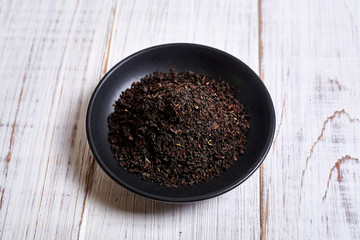 Black dry tea in a clay bowl on a light wooden background.