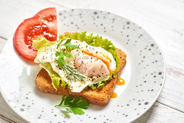 Poached egg, benedict on toasted white bread toast with salad and spices on a plate on a light wooden background.