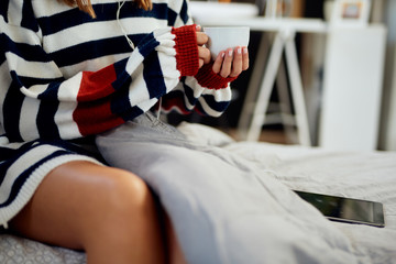 Caucasian woman in striped sweater sitting on bed, listening music over tablet and holding her fresh morning coffee.