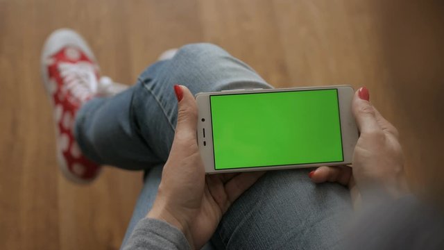 Girl Using Touchscreen Mobile Phone. Young Woman Home Sitting on a Chair with Green Screen Smartphone in Horizontal Mode. Girl Using Smartphone, Browsing Internet, Watching Video Content, Blogs. POV.