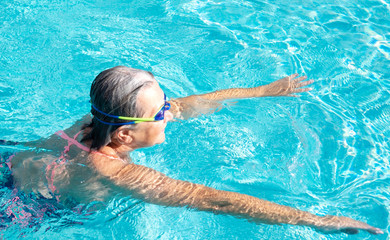 A senior woman with swimming pool goggles swims in the transparent water. Healthy activity for pensioner.