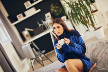 Charming brunette in blue sweater sitting in bedroom in morning and drinking coffee.