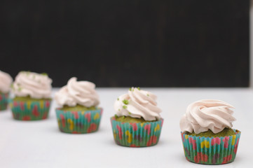 Green cupcakes with cream hat in multi-colored papers on a white table on a dark background. Focus on cake, depth of field effect