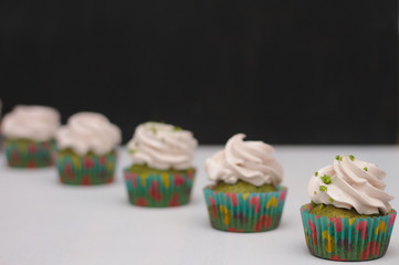 Green cupcakes with cream hat in multi-colored papers on a white table on a dark background. Focus on cake, depth of field effect