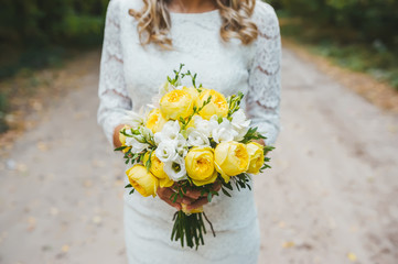A caucasian woman is standing on pathway, holding a bridal bouquet of yellow roses wearing a wedding dress.