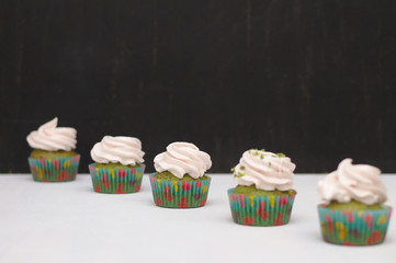 Green cupcakes with cream hat in multi-colored papers on a white table on a dark background. Focus on cake, depth of field effect