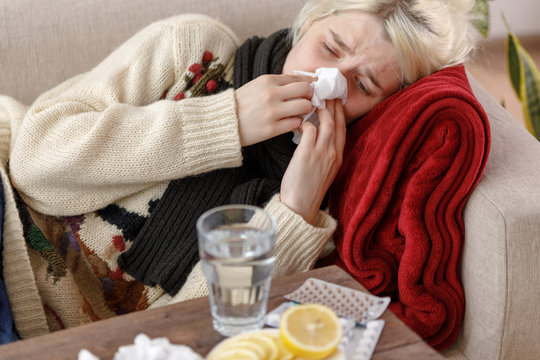 Girl in a sweater sneezes while sitting on a sofa. Colds and flu. The patient caught a cold, feeling sick and sneezing in a paper napkin. An unhealthy girl wiped her nose.