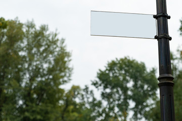 empty white signboard on an iron pole against a light sky and treetops with copy space for your text. blank information sign mock-up