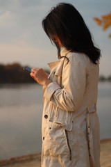 White dark haired girl looks at the cellphone and swipes a finger on the phone screen on a background of an autumn lake