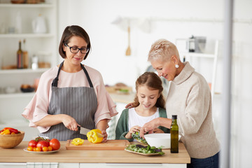 Young mother and grandmother teaching little girl to prepare vegetable salad in the kitchen at home