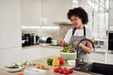 Attractive mixed race woman in apron mixing vegetables in bowl while standing in kitchen at home.