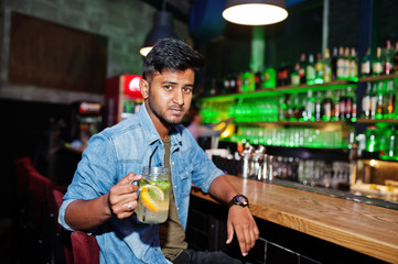Portrait of handsome successful bearded south asian, young indian freelancer in blue jeans shirt sitting in night club against bar counter with cocktail and having a rest.