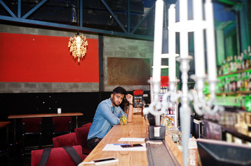 Portrait of handsome successful bearded south asian, young indian freelancer in blue jeans shirt sitting in night club against bar counter with cocktail and having a rest.