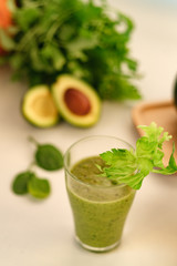 Green smoothie with a stalk of celery in a glass cup. On the table is an avocado.