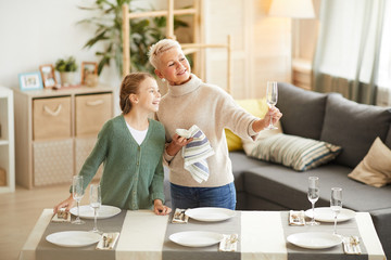 Mature woman wiping the wineglasses and showing them to the little girl while they setting the table together at home