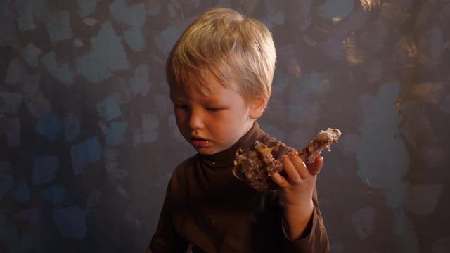 Charming blond boy in dark clothes eats a large piece of meat on the bone.Concept of healthy farm food for children. Strong and healthy country child