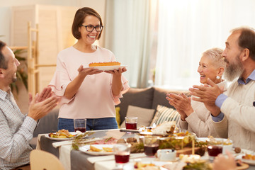 Happy woman with short hair in eyeglasses holding sweet cake and smiling and her family applauding...