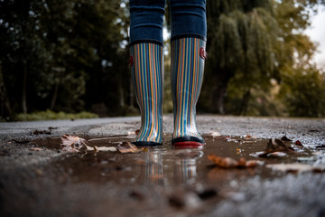 A woman wearing rubber boots jumps into a puddle on a cold autumn day.