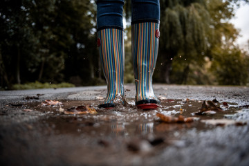A woman wearing rubber boots jumps into a puddle on a cold autumn day.