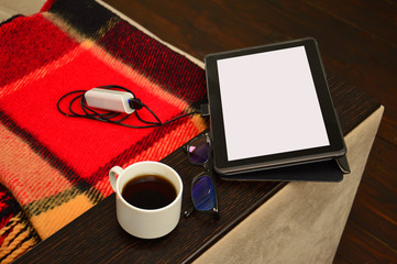 The tablet is connected to the power bank, the gadget is charging. Portable charging, a telephone, glasses and coffee are on the couch.