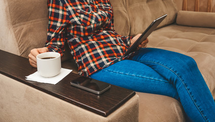 A girl sits on a couch, drinks coffee, holds a tablet and reads the news.