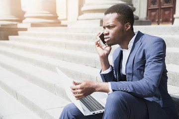 An african young man sitting on staircase holding laptop talking on mobile