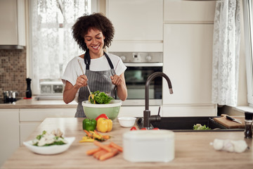 Attractive mixed race woman in apron mixing vegetables in bowl while standing in kitchen at home.