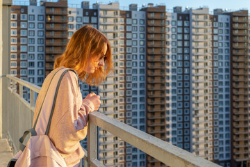 Fototapeta na wymiar Tween redhead girl in pullover, jeans and sunglasses standing on balcony against high-rise multi-storey residential building at sunset. Beautiful look, fashionable city street outfit, teenage fashion