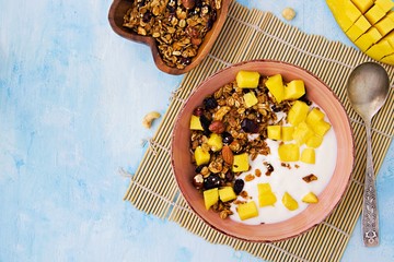 Baked oat granola with nuts, dried fruits, unsweetened natural yogurt and fresh mango in a clay bowl on a light blue background. Tropical breakfast concept. Healthy eating