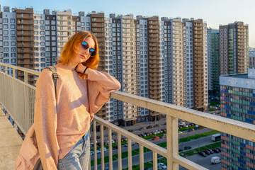 Tween redhead girl in pullover, jeans and sunglasses standing on balcony against high-rise multi-storey residential building at sunset. Beautiful look, fashionable city street outfit, teenage fashion