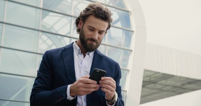 Handsome Businessman browsing his Smartphone near modern Office Building. Attractive Man is smiling and wearing Smart-casual Style. Social Networking. Luxury Lifestyle. Smartphones. Apps. - Powered by Adobe