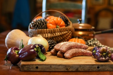 Selective focus still life of preparation of German bratwurst purple peppers and onions on wooden cutting board with a large knife, with blurred basket of pumpkins and pine cones in background 