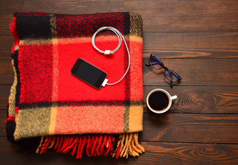 The phone is connected to the power bank, the gadget is charging. Portable charging, a telephone, glasses and coffee are on a wooden table.
