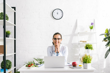 cheerful nutritionist in white coat near vegetables and laptop - obrazy, fototapety, plakaty