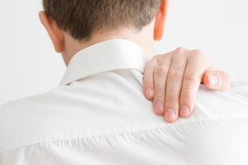 Young man in white shirt massaging his shoulder with hand. Pain from strain of sedentary long work, overwork and incorrect posture. Inactive lifestyle. Back view. Close up.