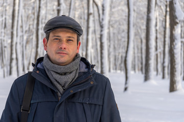 Portrait of handsome middle-aged man walking in winter snowy park or forest. Attractive man in jacket, scarf and cap looking at camera. Winter mood, authentic lifestyle concept, stylish male outfit