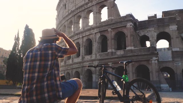 Happy young man tourist wearing shirt and hat with bike sitting and taking pictures with vintage camera at colosseum in Rome, Italy at sunrise.