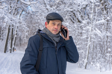 Fototapeta na wymiar Handsome middle-aged man walking in winter snowy park or forest. Attractive man in jacket, scarf and cap talking on mobile phone. Winter mood, authentic lifestyle concept, stylish male outfit